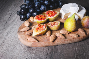 Still life cheese plate on a wooden cutting board lies sliced cheese with mold almonds in the shell and sliced figs next to a bunch of black grapes. Delicious and healthy food concept