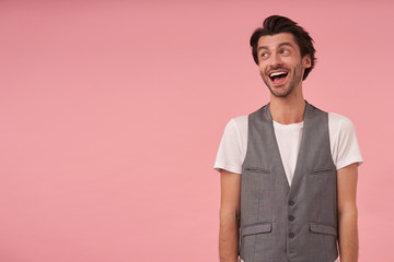 Happy young dark haired man wearing grey waistcoat and white t-shirt, standing over pink background with hands down, looking aside with wide smile, positive emotions concept