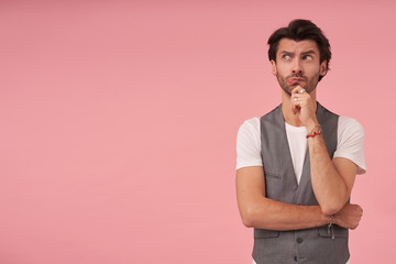 Young pensive dark haired man in grey waistcoat and white t-shirt standing over pink background, looking aside with thoughtful face and holding chin with his hand, frowning and contracting forehead
