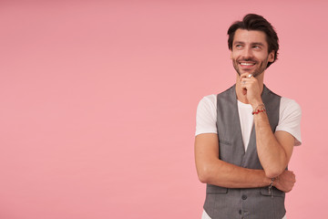 Indoor shot of cheerful bearded male with trendy haircut posing over pink background, wearing grey waistcoat and white t-shirt, holding his chin with hand and looking aside with charming smile