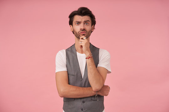 Good looking dark haired male with beard standing over pink background in grey waistcoat and white t-shirt, holding chin with hand and contracting forehead with raised eyebrow