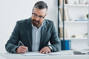 handsome businessman in formal wear and glasses writing with pen in office