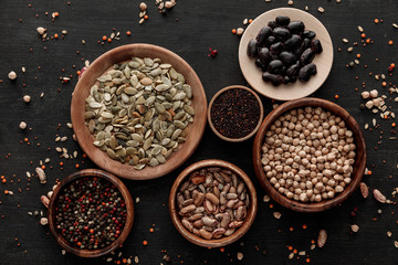 top view of wooden bowls and plates with raw beans, chickpea, quinoa, pumpkin seeds and peppercorns on dark surface with scattered grains