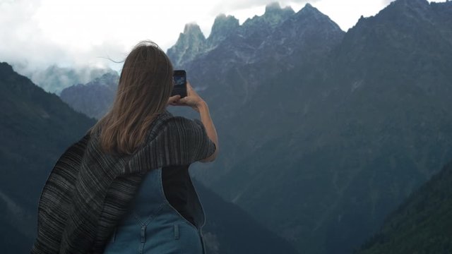 Young attractive girl traveler-blogger makes a photo of large mountains on a smartphone. Dressed in warm clothes and a plaid. Walking along a mountain river. Tetnuldi Mountain, Svaneti, Georgia.