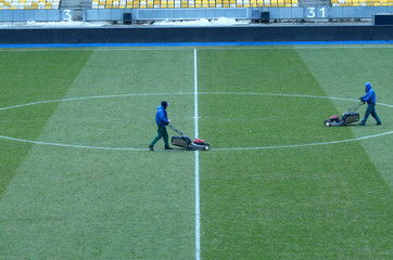 Workers mowing lawn on a soccerl field,  empty seats of stands on a background