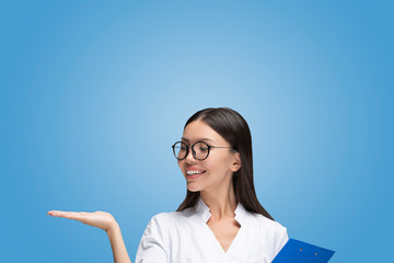 A beautiful young Asian woman doctor is presenting with her hand in front of isolated and easy to remove blue background. Communicate about a healthy or medical option, showcase a product or icon