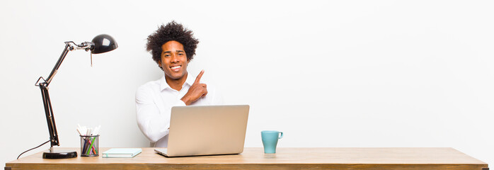 young black businessman smiling cheerfully, feeling happy and pointing to the side and upwards,...