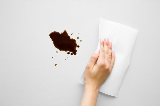 Woman's Hand Cleaning Fresh Spilled Dark Beverage From Gray Desk. Coffee Stain Simple Removing With White Paper Napkin. Cleanup. Close Up.