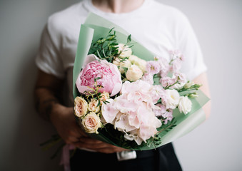 Very nice young man in a white t-shirt holding a beautiful blossoming flower bouquet of fresh peony, hydrangea, roses, mattiola in tender pink colours on the grey wall background 