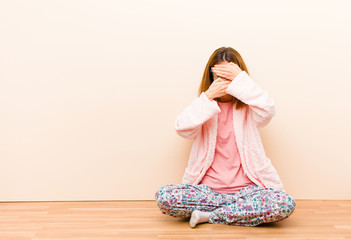 young woman wearing pajamas sitting at home covering face with both hands saying no to the camera!...