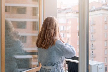 Blonde woman in bathrobe drinking her morning coffee or tea on a downtown balcony. Woman enjoying sunrise and downtown city view from a balcony.