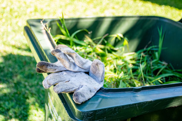 Green bin container filled with garden waste. Dirty gardening gloves. Spring clean up in the garden. Recycling garbage for a better environment.