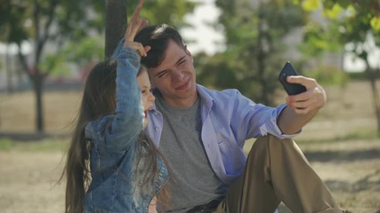 Teenage boy with his small sister taking funny playful selfie photoes on smartphone sitting in park sunny autumn day