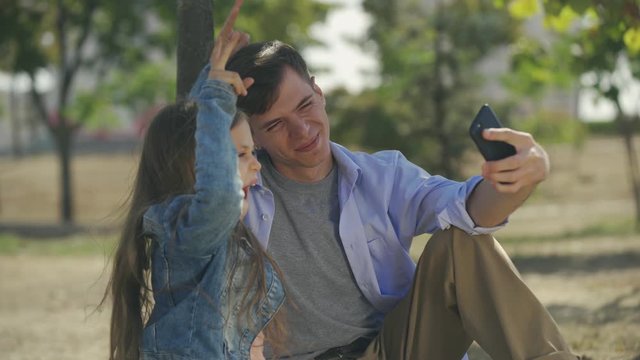 Teenage boy with his small sister taking funny playful selfie photoes on smartphone sitting in park sunny autumn day