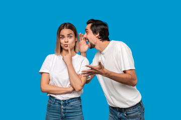 Bearded man with mustaches in white t-shirt tells secret to a young blond girl in white t-shirts and blue jeans standing isolated over blue background. Concept of secret telling.