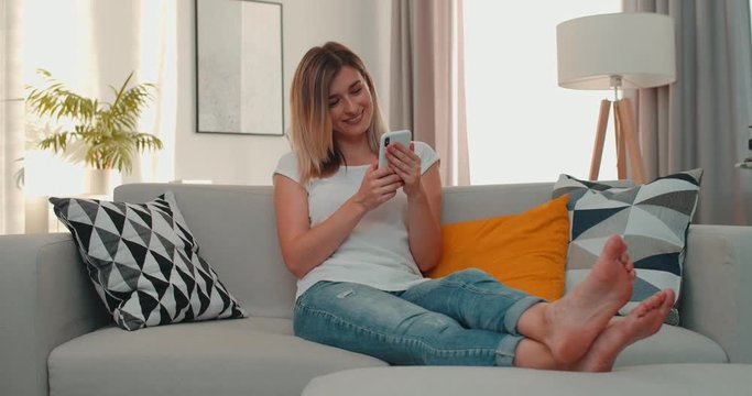 Good looking Caucasian young woman in th ewhite T-shirt and blue jeans sitting and resting on the sofa at home while scrolling photos on the smartphone and looking at the screen with a smile.