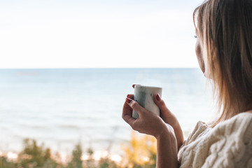 Side view portrait of a woman drinking coffee and looking outdoors through a window of an hotel room or home with the sea in the background