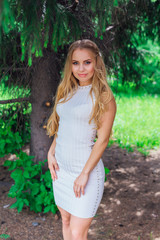 Portrait of a charming blond woman wearing beautiful white dress standing under fur tree.