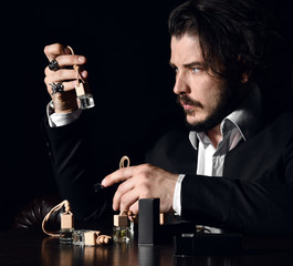 Brutal bearded man with modern hairstyle perfumer examines small bottles of perfume fragrance, holding one on dark