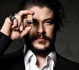 Brutal man with beard and modern hairstyle wearing business classic suit shows small fragrance bottle
