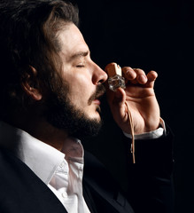 Brutal bearded man with modern hairstyle in official suit and white shirt smells small perfume bottle on dark