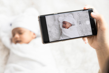 Mother 's hand holding smartphone use to taking photo cute newborn baby for memories and pose in...