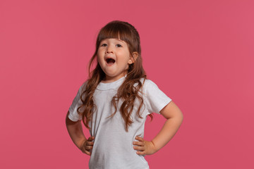 Close-up portrait of a little brunette girl dressed in a white t-shirt posing against a pink studio background. Sincere emotions concept.