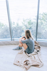 Sad slim woman sitting on a warm floor in a grey dress near the large window in light and hugging her knees. Autumn mood, warmth and comfort.