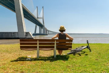 Papier Peint Lavable Pont Vasco da Gama Young woman resting on a bench after biking while is looking at Vasco da Gama bridge in Lisbon.