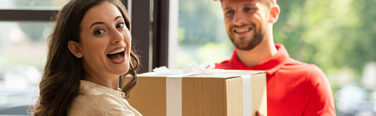 panoramic shot of happy woman receiving carton box from cheerful delivery man