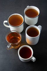Top view of various cups, mugs with hot tea drink on dark background, copy space. Tea time or tea brake. Autumn beverage. Toned image with tea cups.