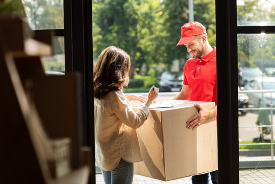 selective focus of happy delivery man holding box near woman with pen