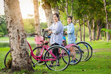 Happiness asian mother and daughter standing near their bicycle on green grass in city park, smiling child girl and happy woman is resting on holiday and looking at camera, health lifestyle concept