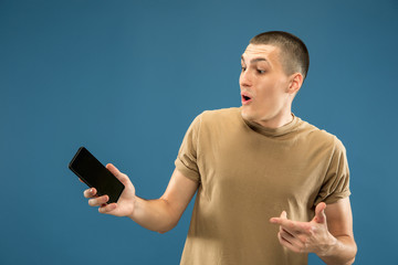Caucasian young man's half-length portrait on blue studio background. Beautiful male model in shirt. Concept of human emotions, facial expression, sales, ad. Showing phone screen, looks astonished.