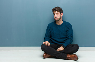 young handsome man sitting on floor feeling sad and stressed, upset because of a bad surprise, with...