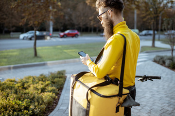 Young bearded courier delivering food in thermal bag on a bicycle, checking order with a smart...