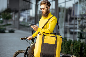 Fototapeta na wymiar Young bearded courier delivering food in thermal bag on a bicycle, checking order with a smart phone while standing on the street outdoors