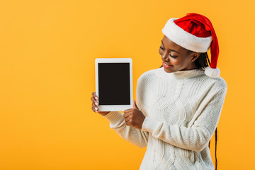 African American woman in winter sweater and Santa hat holding digital tablet with blank screen isolated on yellow