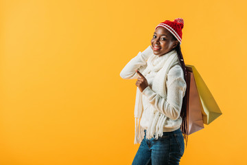 side view of African American woman in winter outfit holding shopping bags and pointing with finger isolated on yellow