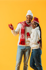 Fototapeta premium African American man in cozy winter outfit hugging woman and pointing with hand isolated on yellow