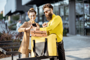 Male courier delivering pizza to a young businesswoman, client signing on a smart phone near the office building. Delivery service concept