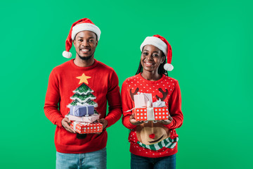 African American couple in red Christmas sweaters and Santa hats holding presents and looking at camera isolated on green