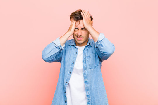 young handsome man feeling stressed and anxious, depressed and frustrated with a headache, raising both hands to head against pink background