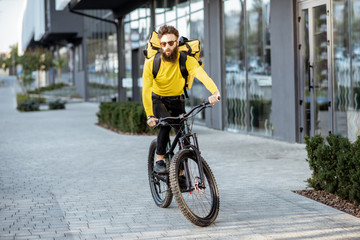 Plakat Young bearded courier delivering food with a yellow thermal backpack, riding a bicycle in the city. Food delivery service concept