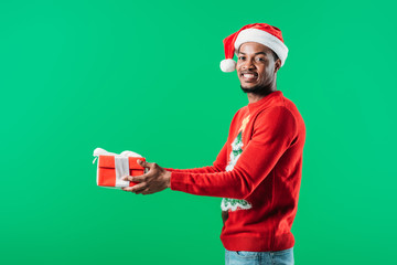 side view of African American man in Christmas sweater and Santa hat holding red gift box looking at camera isolated on green