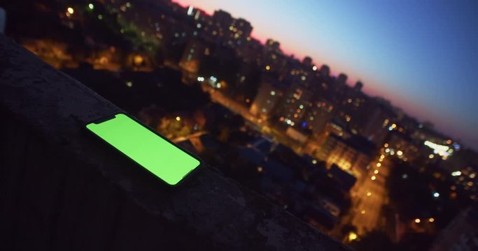 Mockup greenscreen smartphone modern gadget lying on egde of viewpoint above stunning evening cityscape. Close-up.