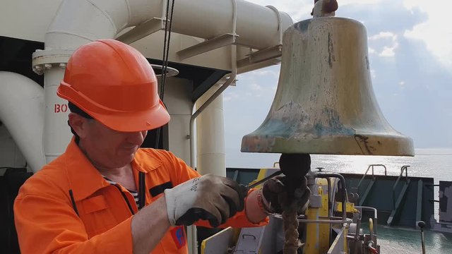 bosun in orange jumpsuit helmet and white gloves cleans large yellow noon bell on oil tanker deck under blue sky closeup