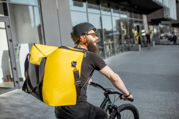 Fototapeta na wymiar Young bearded courier delivering food with a yellow thermal backpack, riding a bicycle in the city. Food delivery service concept
