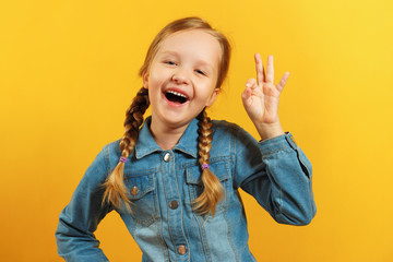 Little girl in a denim shirt on a yellow background. The child shows an OK sign. The concept of...