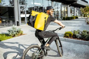 Young bearded courier delivering food with a yellow thermal backpack, riding a bicycle in the city....